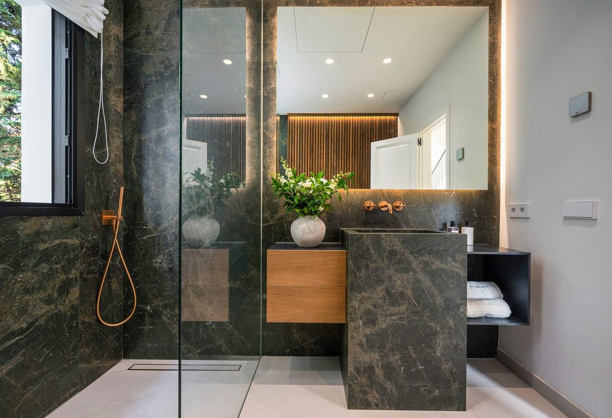 Dark marble bathroom with shower, single stone sink, wooden vanity, and large mirror with backlighting.