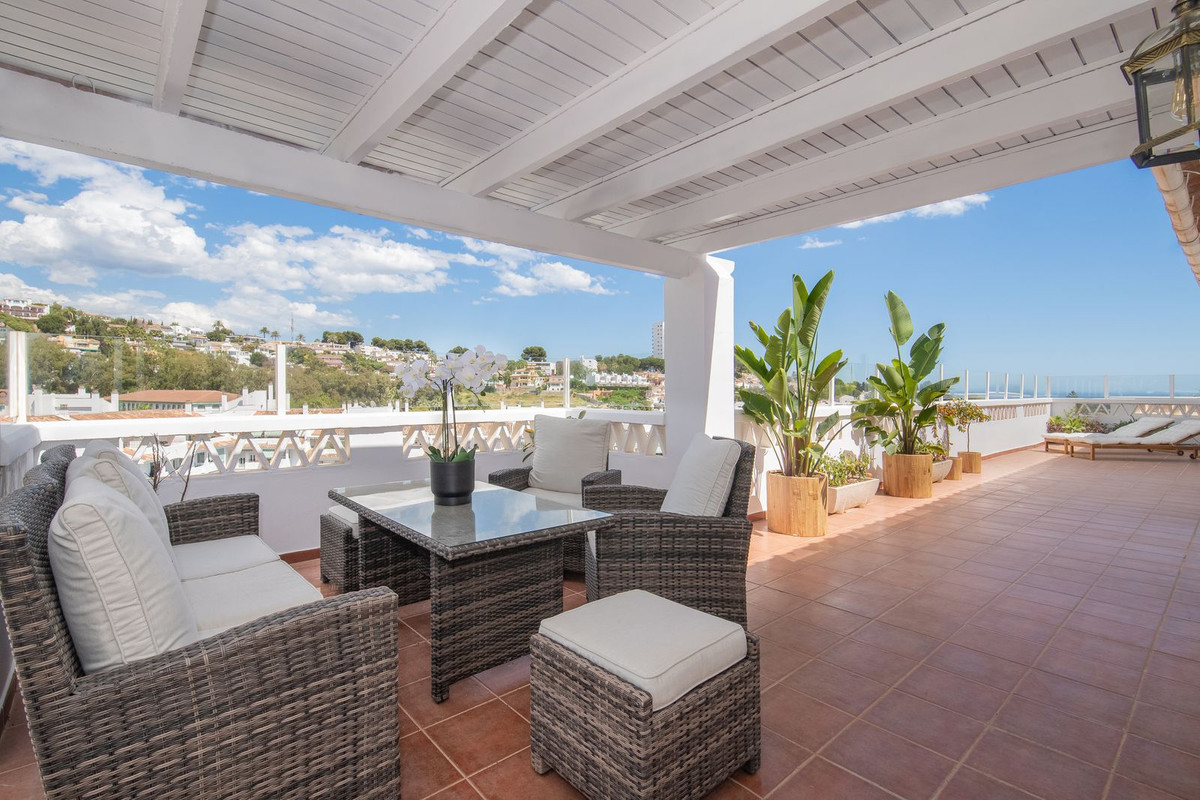 Rooftop terrace with woven outdoor seating, a glass table, potted plants, and a partial town view.