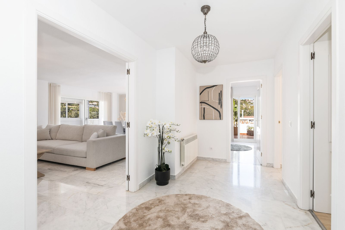 White hallway with marble floor, a round rug, and an orchid plant, leading to a living area.