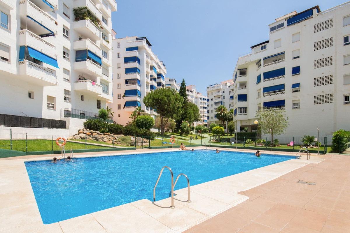 Outdoor community pool with swimmers, surrounded by white apartment buildings featuring blue awnings and lush landscapin