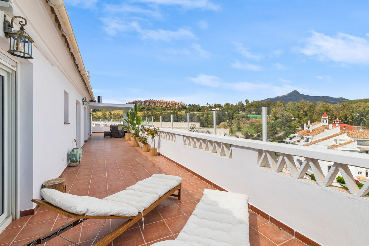 Spacious rooftop terrace with two wooden lounge chairs, white railing, and a view of mountains and town.
