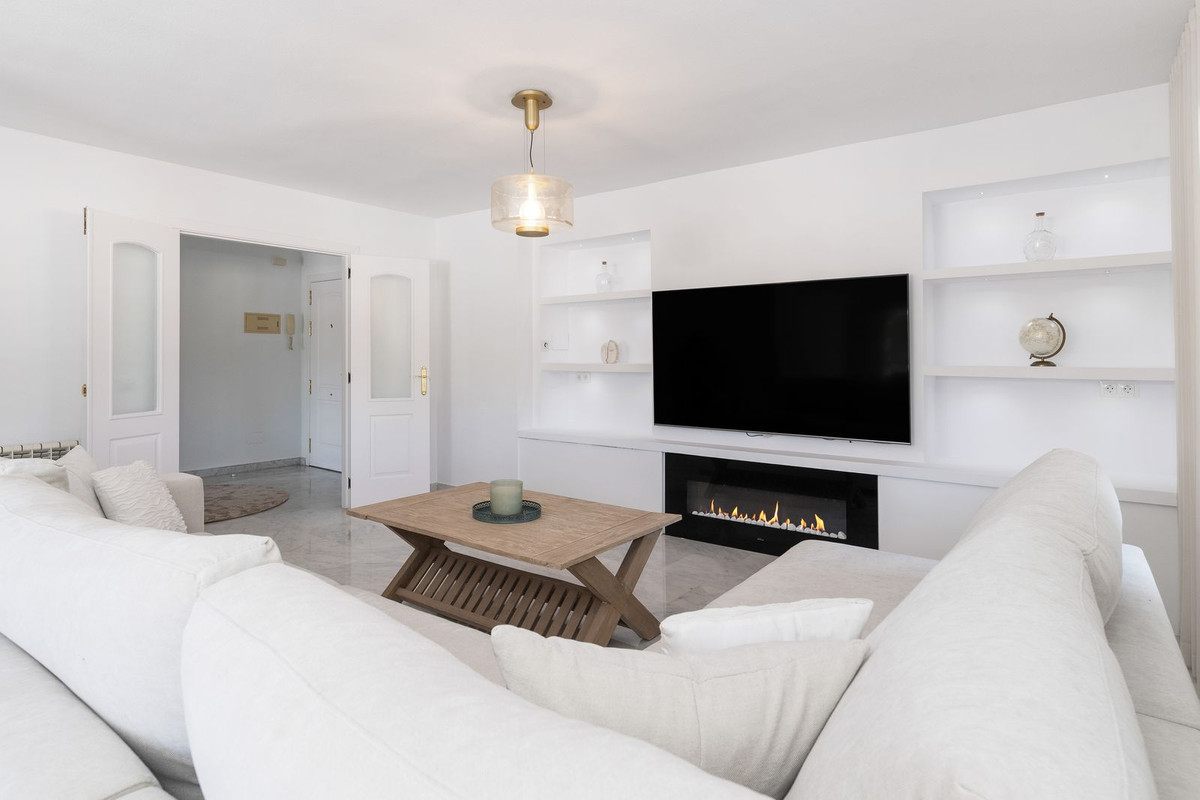 Living room showing a white L-shaped sofa, a TV above a fireplace, and open doors to an entrance hall.