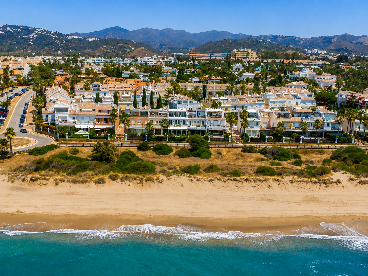 Aerial view of a coastal town with white buildings, palm trees, sandy beach, and turquoise ocean.