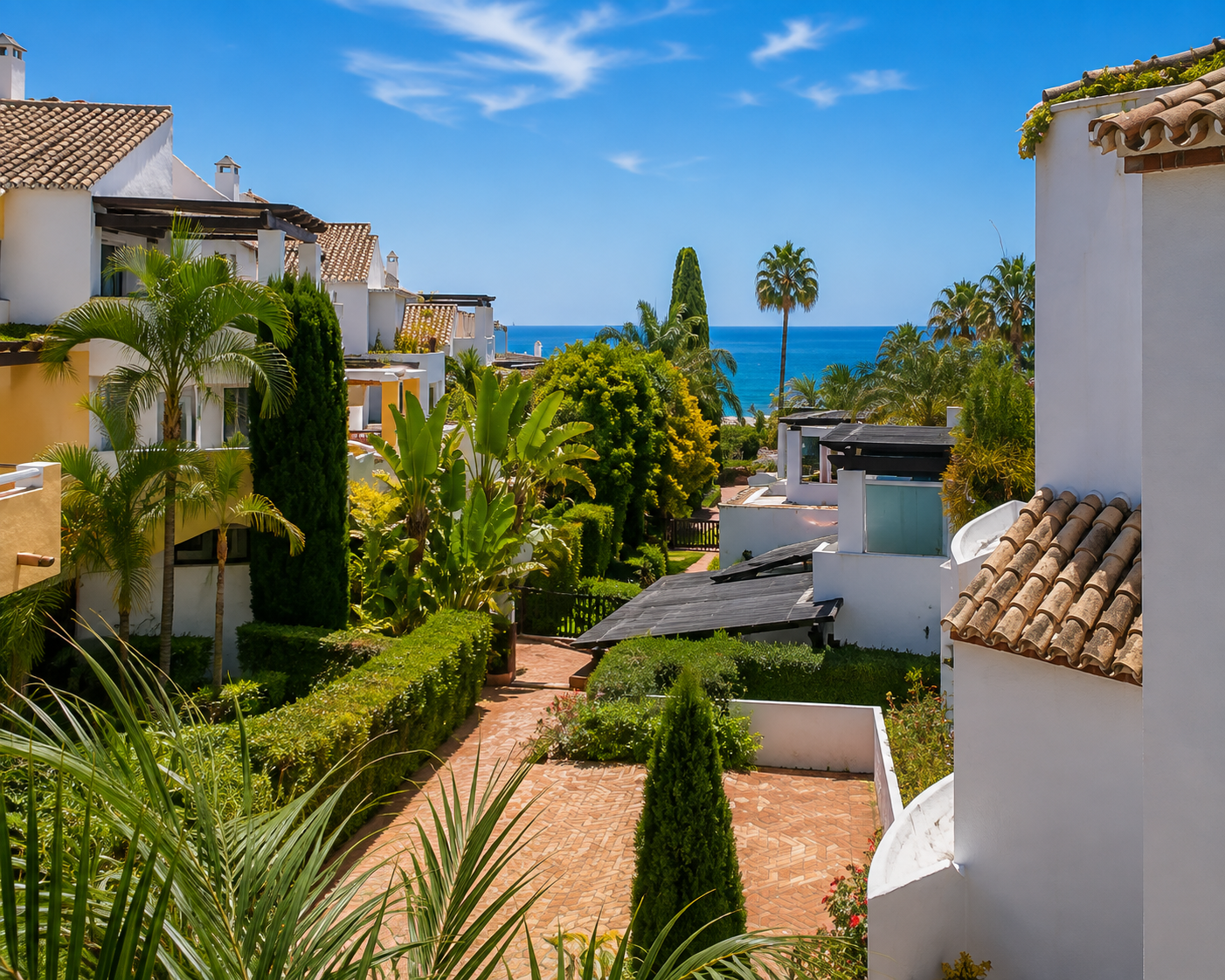 Overhead view of white buildings with terracotta roofs, lush gardens, and a pathway leading to the blue ocean.
