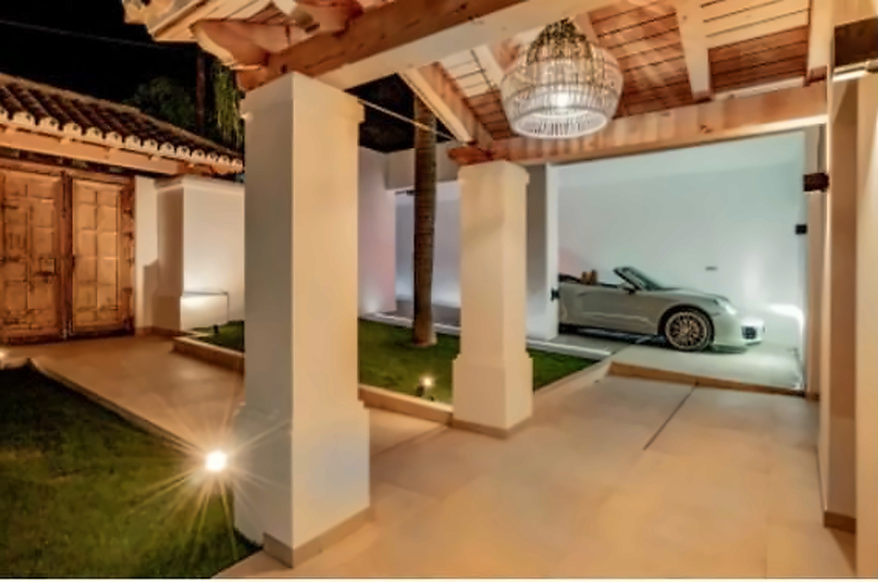 Night view of a covered driveway with a parked silver convertible, lit pillars, and a circular light fixture.