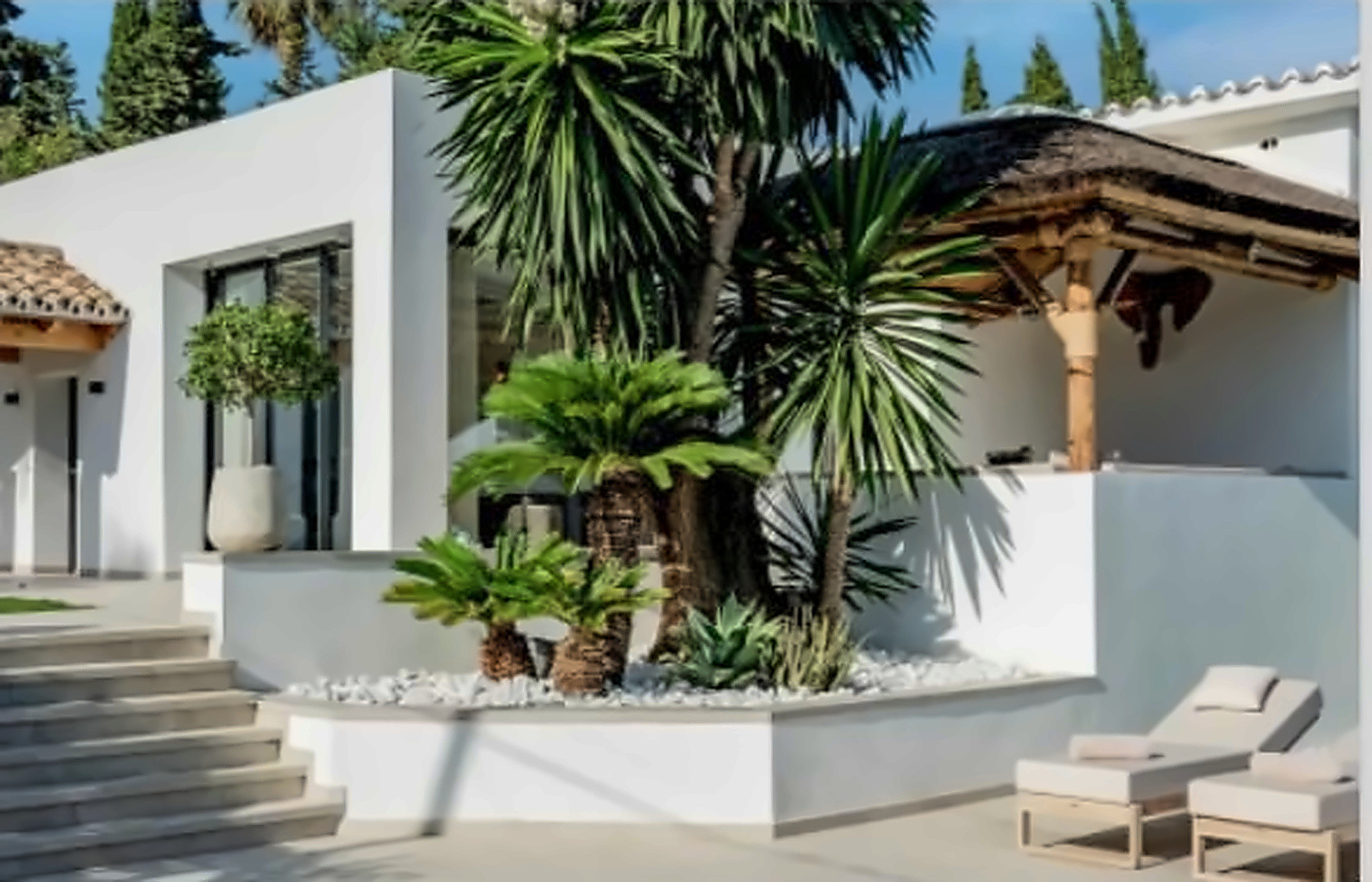 Exterior view of a white villa with palm trees, a thatched pergola, and sun loungers near steps.