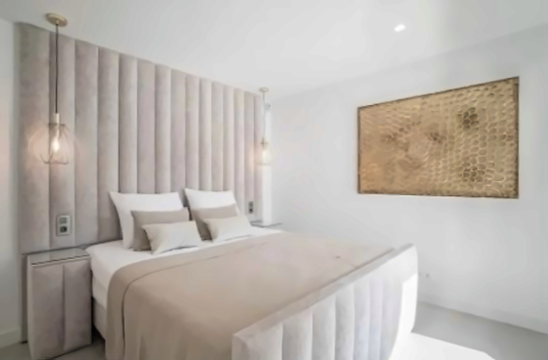 Bedroom with a padded headboard, hanging pendant lights, a bed with neutral linens, and gold wall art.