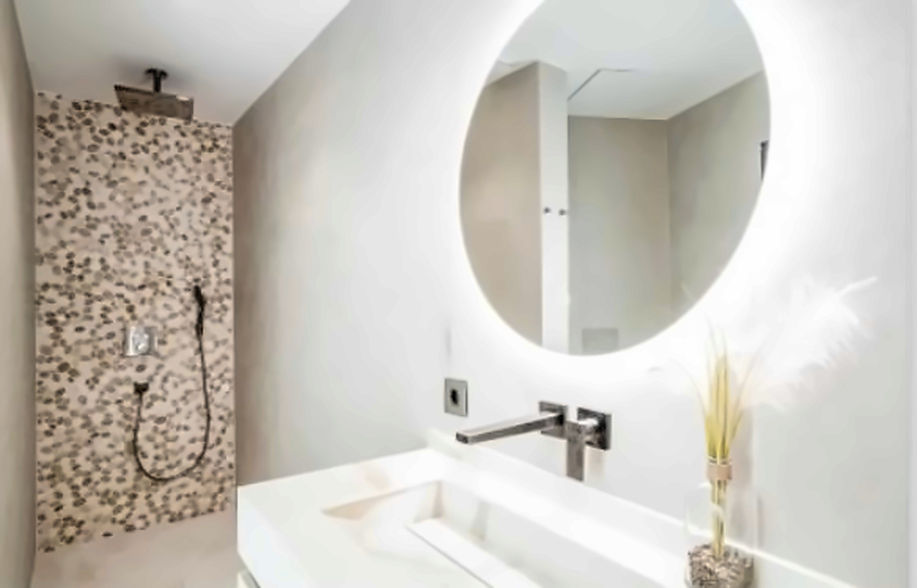 Bathroom vanity with white trough sink, backlit round mirror, and tiled walk-in shower.