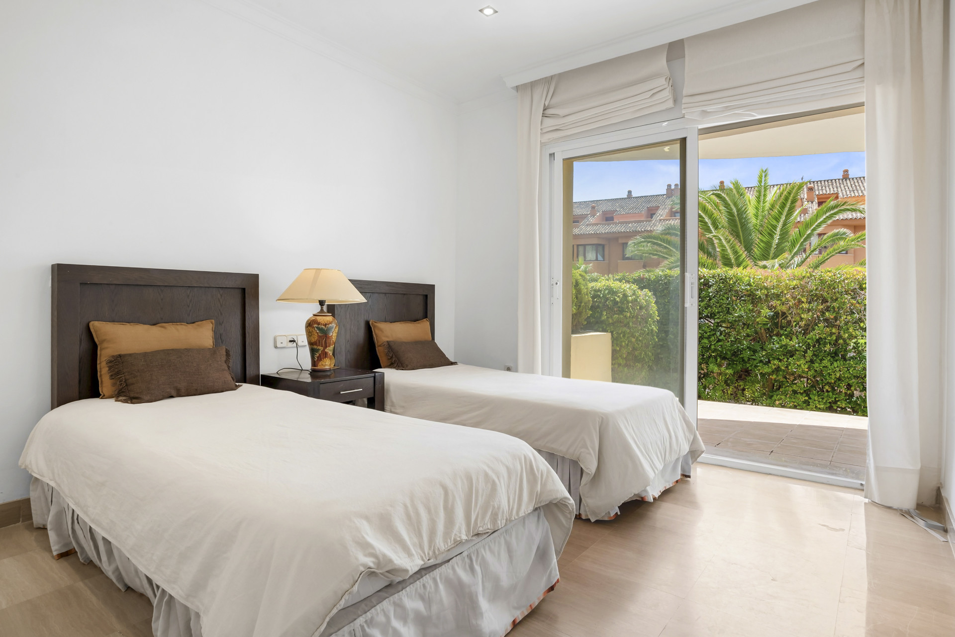Bedroom with two single beds, dark wood headboards, and glass doors leading to a patio with foliage.