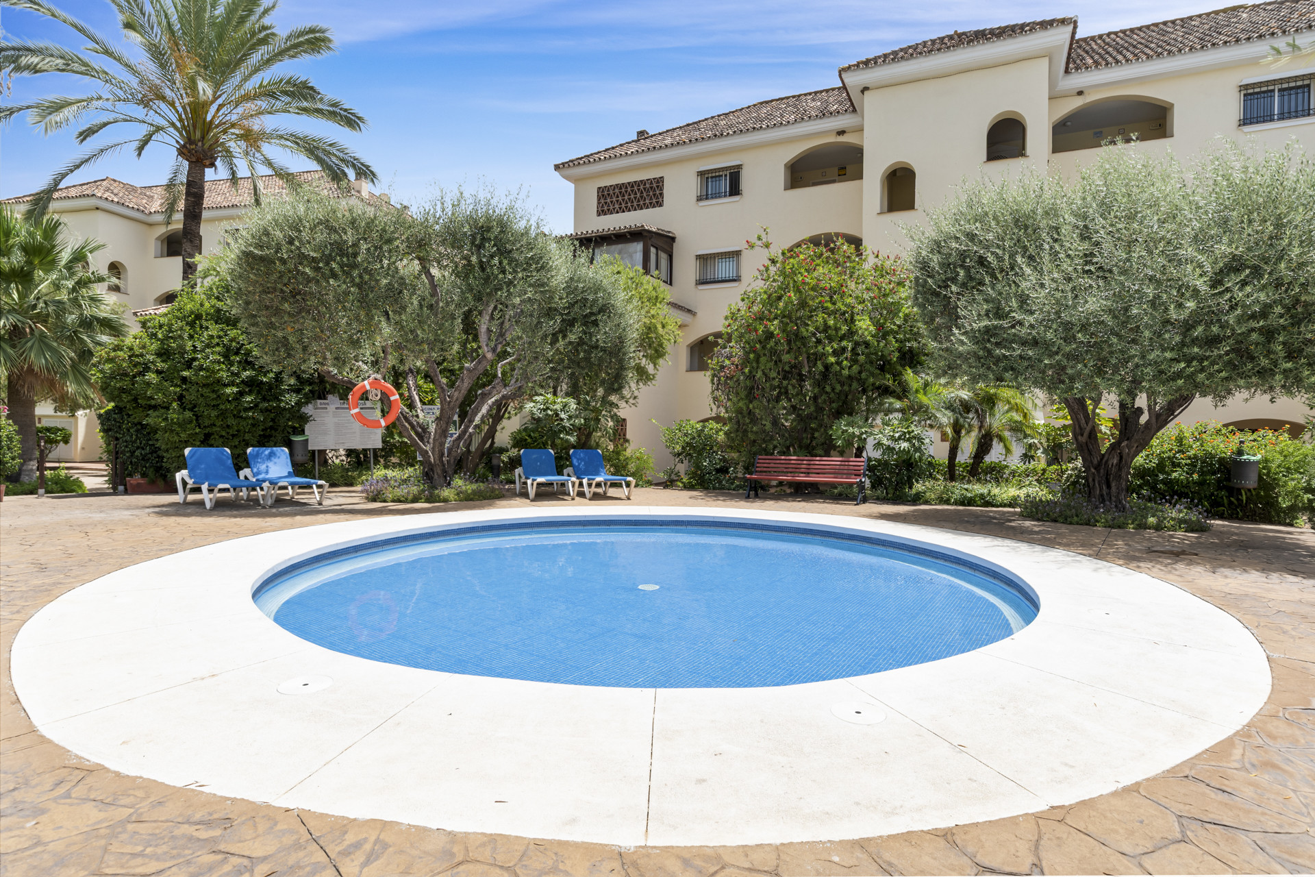Small circular pool on patterned concrete, flanked by mature trees, lounge chairs, and apartment complex buildings.