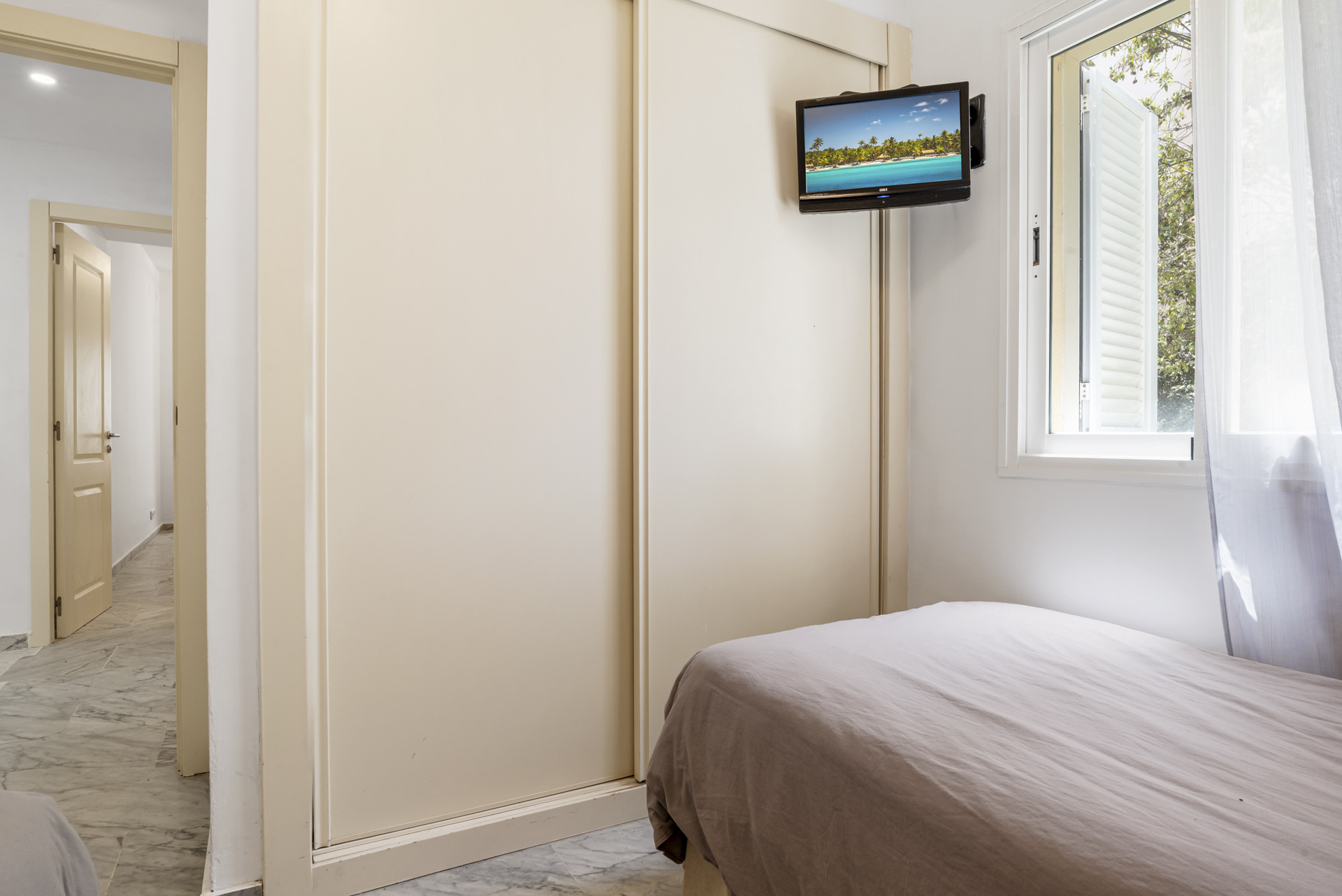 Bedroom corner with a single bed, closed sliding wardrobe, wall-mounted TV, and window.
