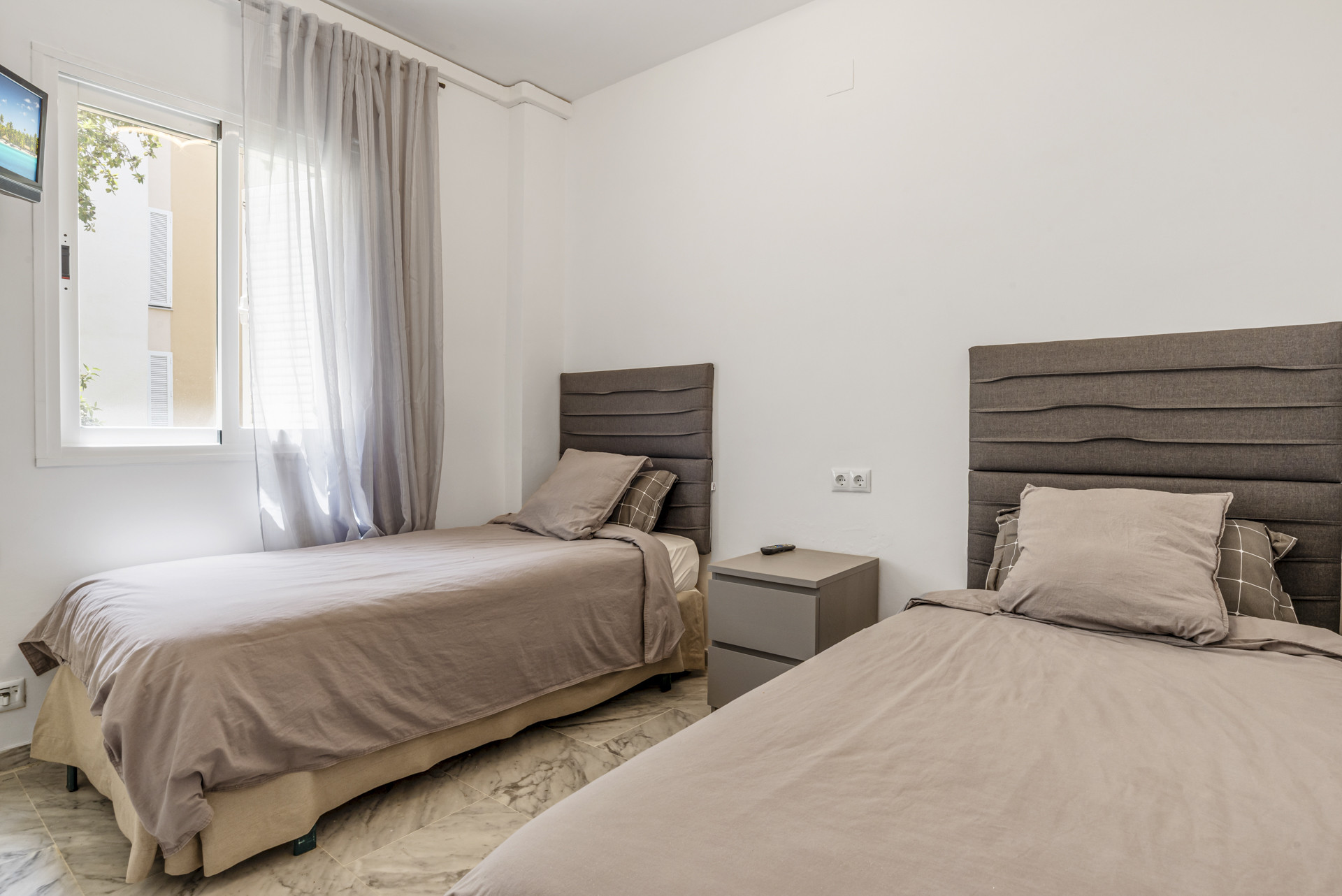 Bedroom with two single beds, gray headboards, and a window with sheer curtains.
