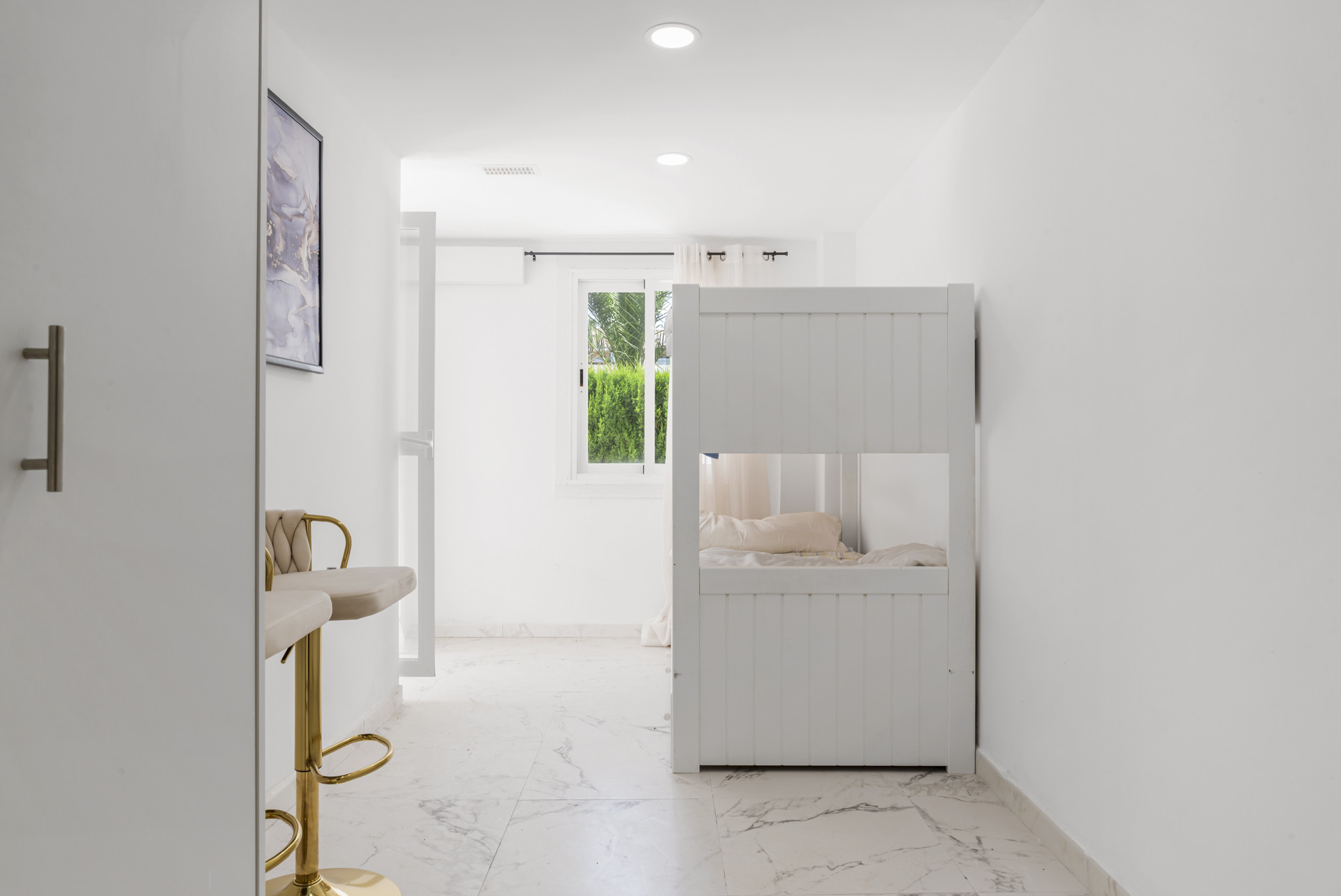 White hallway with bunk beds on the right, a gold bar stool on the left, and a window at the end.