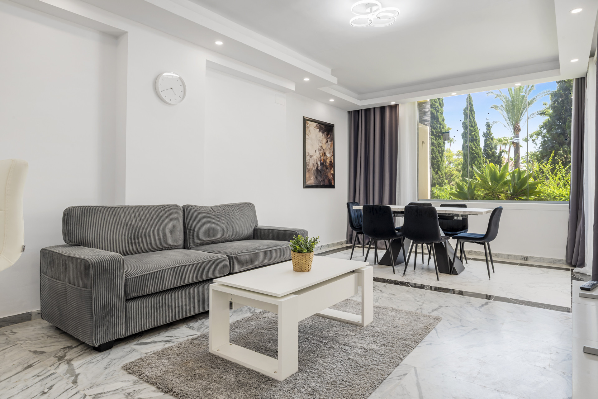 Living and dining area with gray sofa, white coffee table, and dining table by a large window.