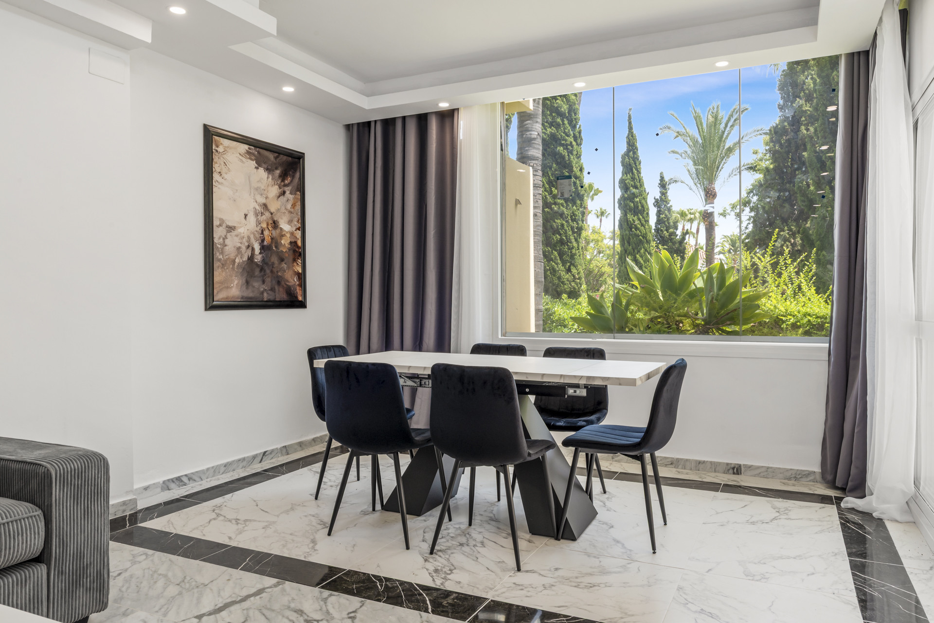 Dining area with black table and six chairs, large window view of tropical trees and landscaping.