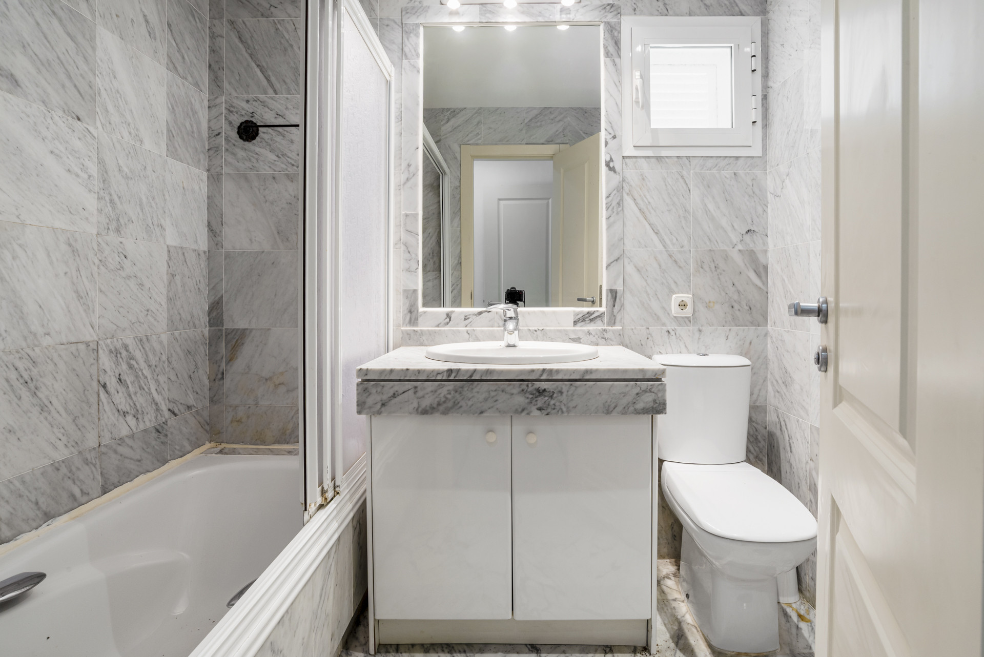 Bathroom with marble tiled walls, bathtub, single vanity, mirror with lights, and white toilet.