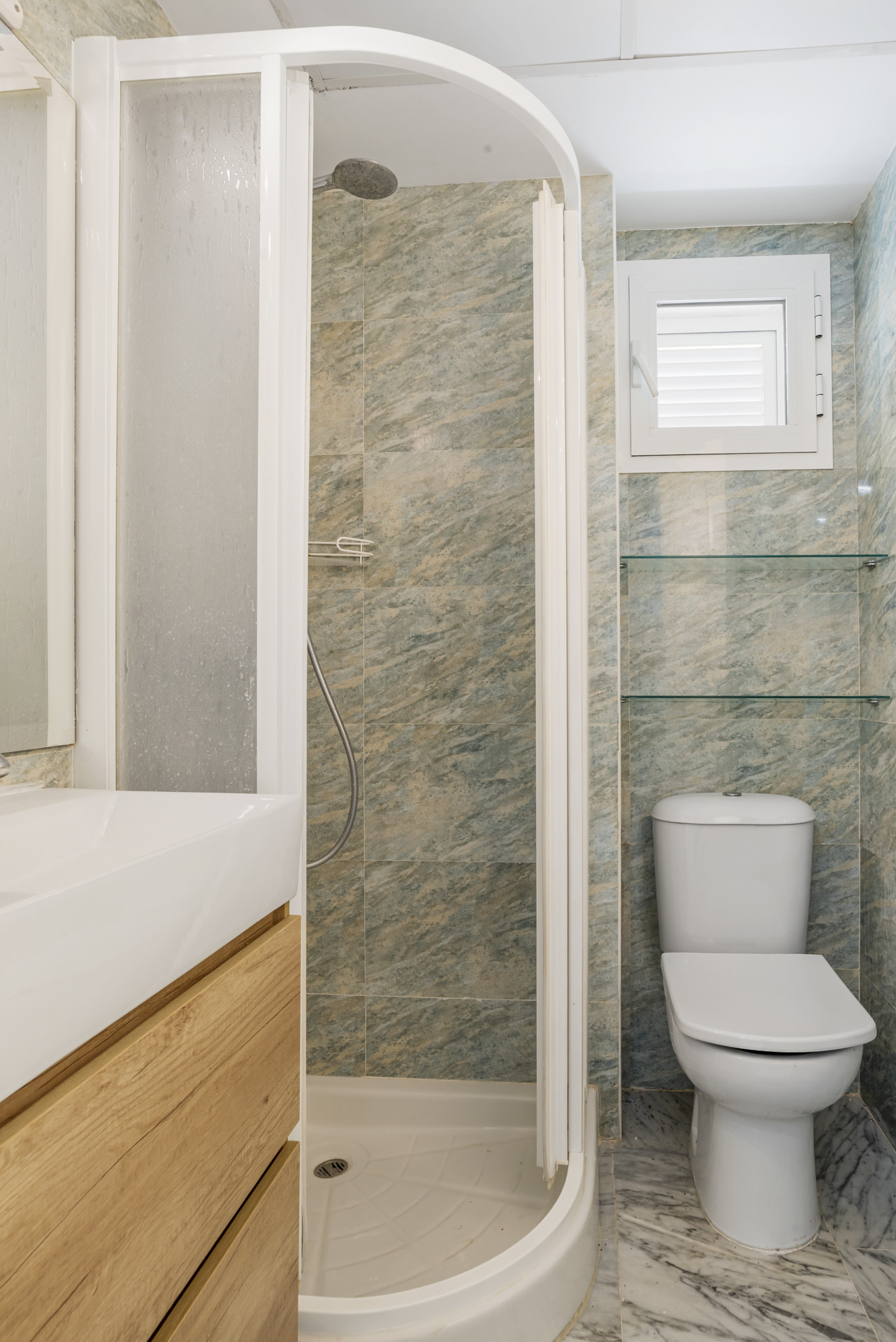Small bathroom showing a white toilet, a rounded shower stall, and a small window with blinds.