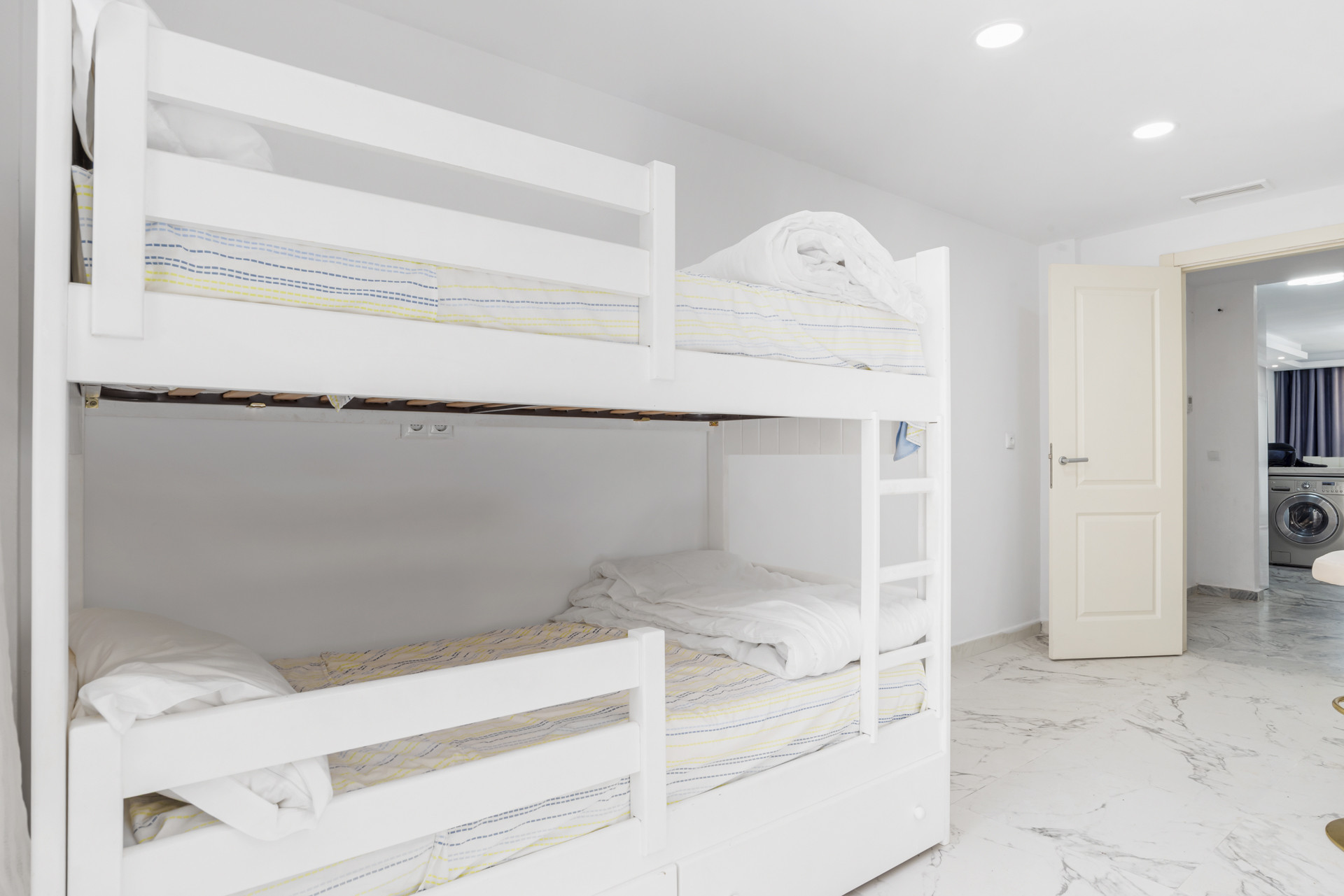 White bedroom with white bunk beds, a light patterned duvet, and an open doorway to another room.