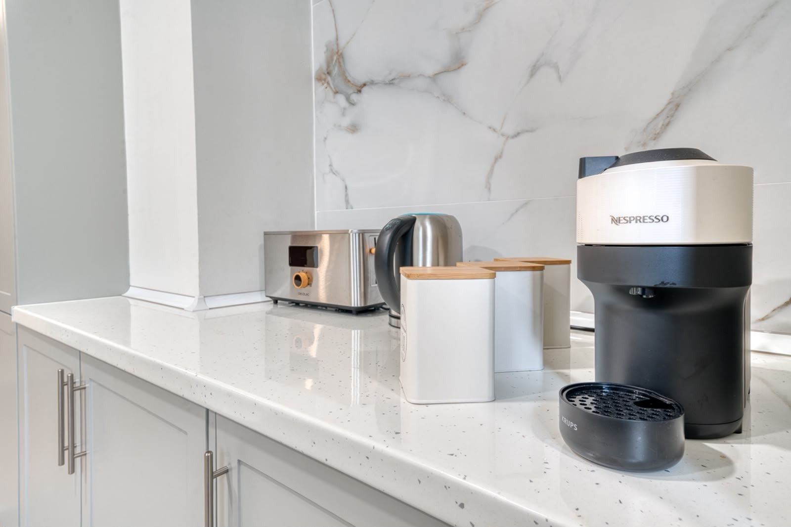 Kitchen countertop with a coffee maker, toaster, kettle, and storage canisters against a tiled backsplash.
