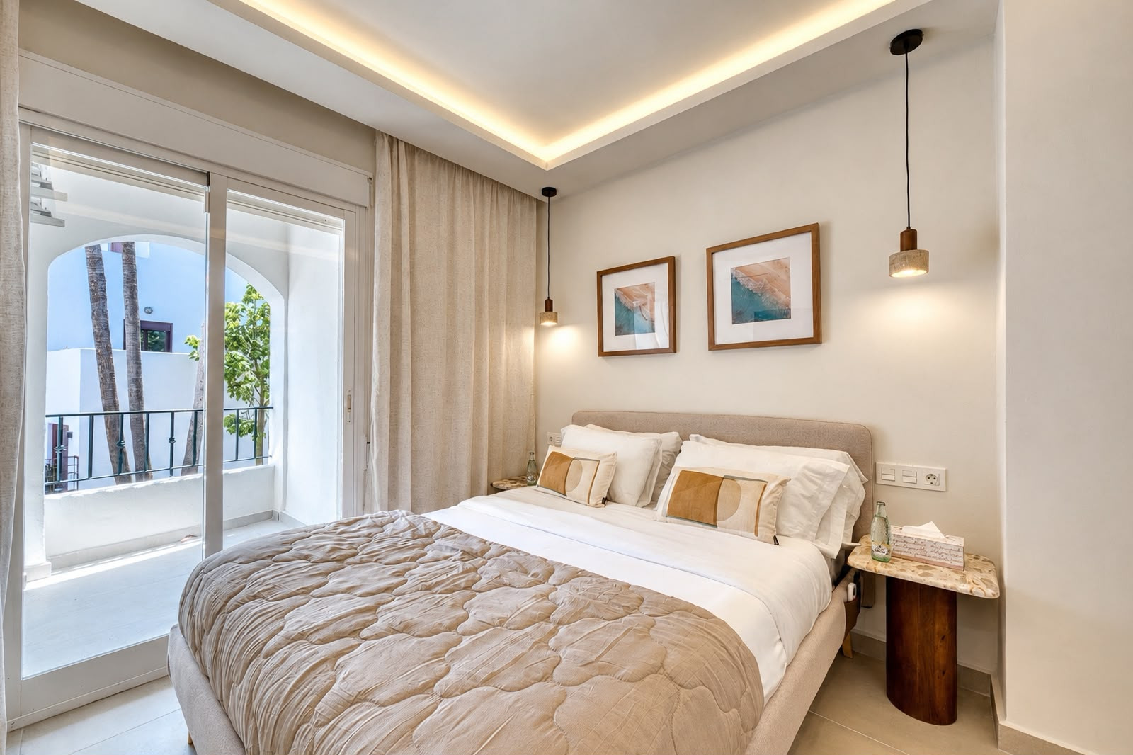 Bedroom with a large bed, two framed artworks, suspended pendant lights, and a sliding door to a balcony.