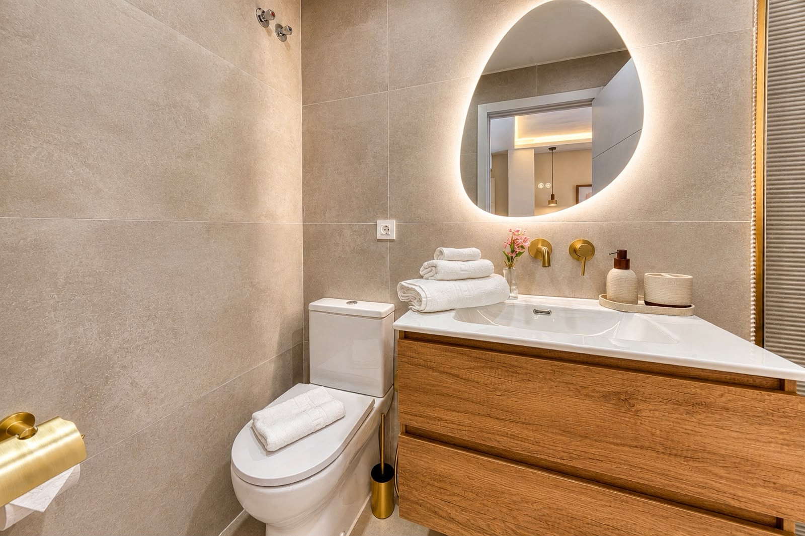 Modern bathroom corner with toilet, wooden vanity, single sink, backlit irregular mirror, and gold fixtures.