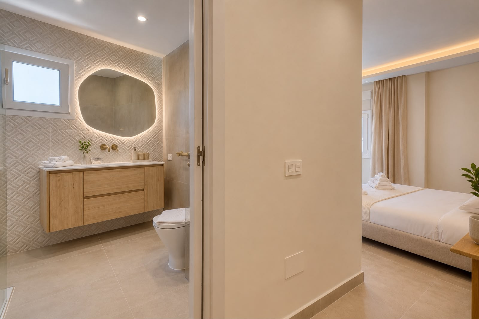 View from hallway into modern bathroom with illuminated mirror and bedroom with beige bed and lighting.