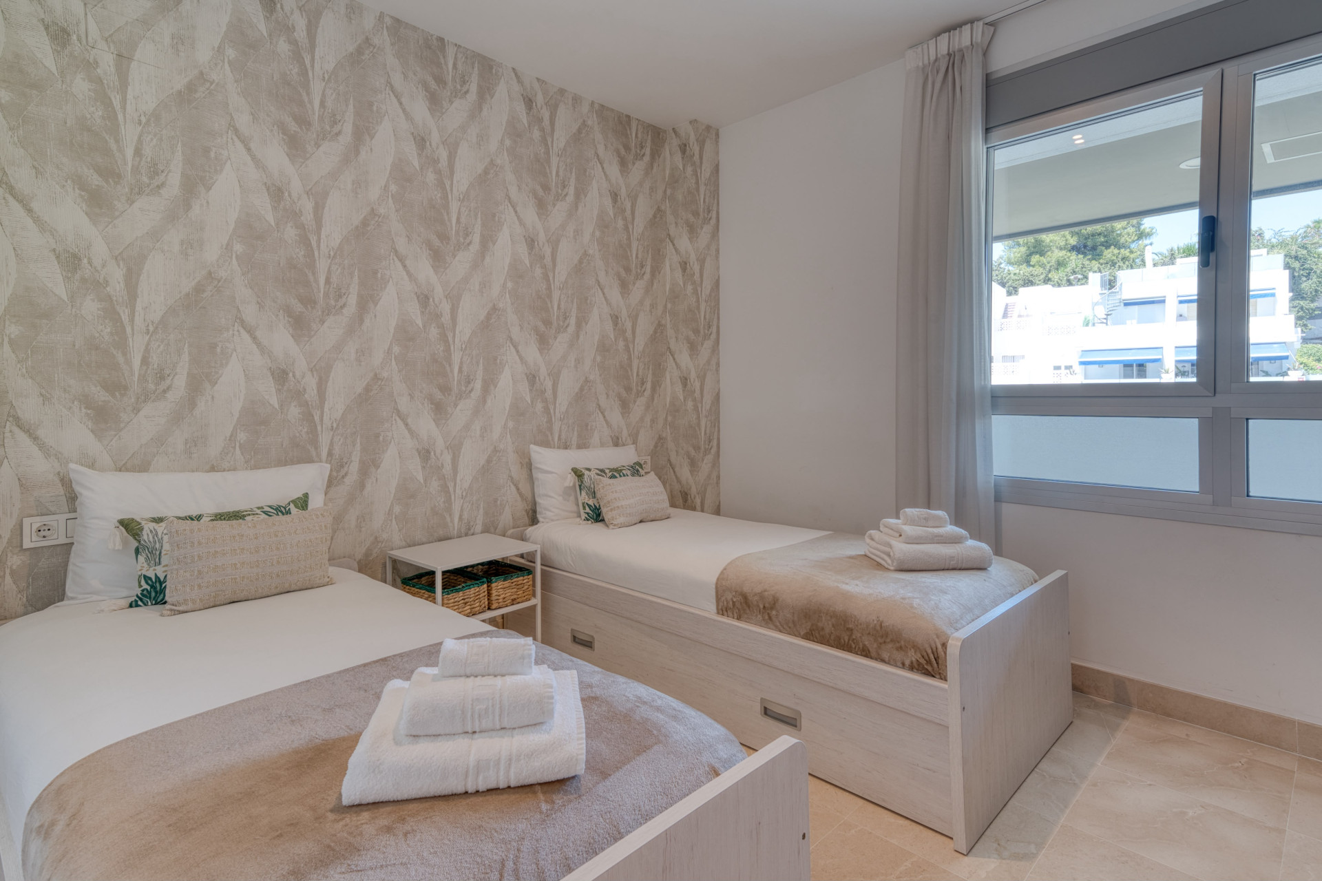 Bedroom with two single beds, leaf-patterned wallpaper, and a window overlooking a residential area.