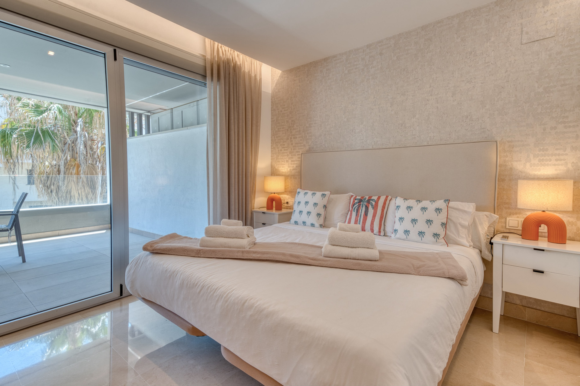 Bedroom with large bed, two nightstands, and a glass sliding door to a balcony with palm trees.