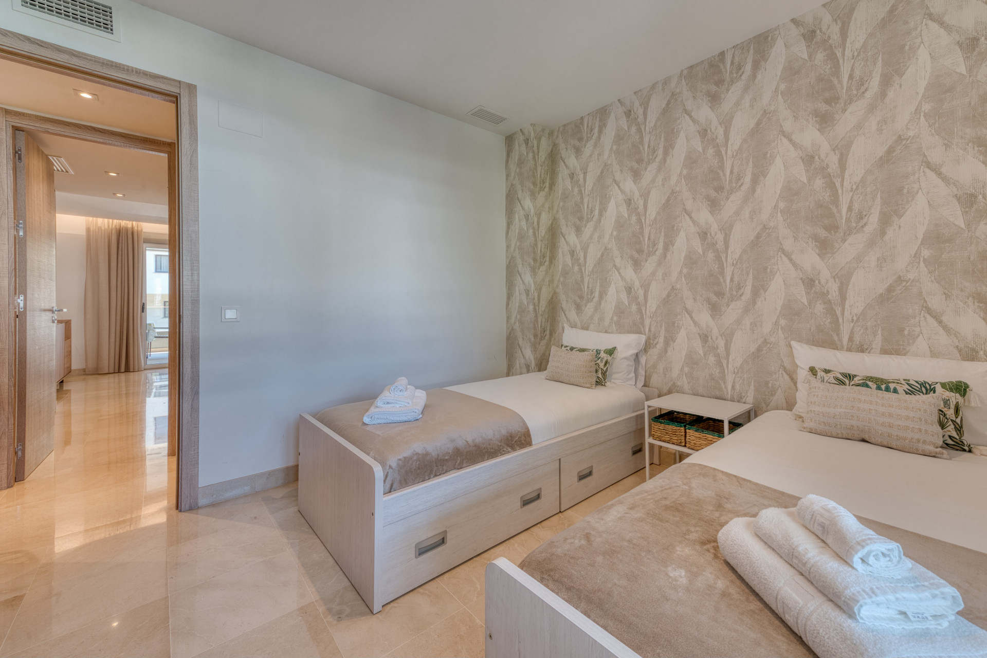 Bedroom with two single beds, patterned wallpaper, and an open doorway revealing a bright hallway.