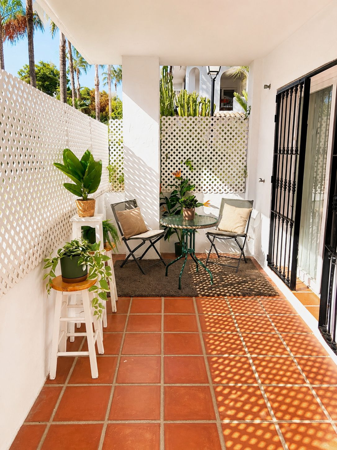 Wider view of the outdoor patio with terracotta tiles, white lattice fencing, and a small round table with two chairs.