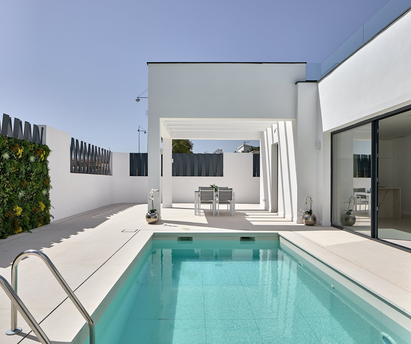 Exterior view of a modern white building with a rectangular pool and an outdoor dining area under a pergola.
