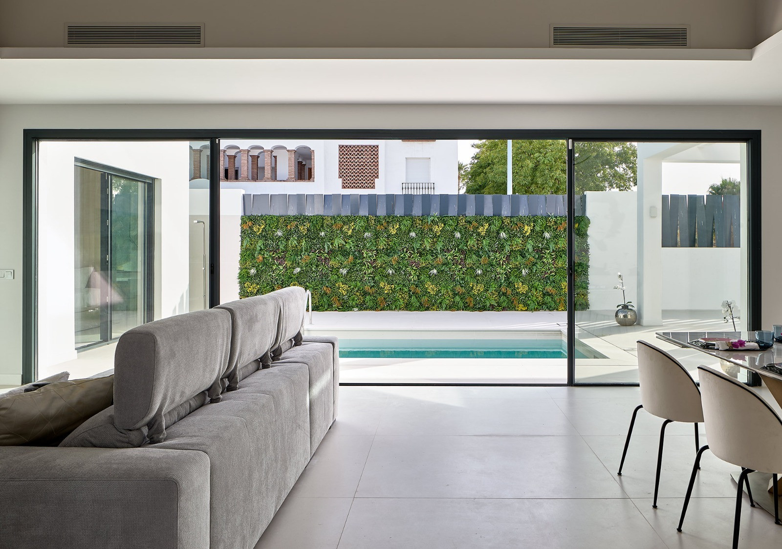 Living room view from behind a gray sofa, looking out through glass doors at a green wall and swimming pool.