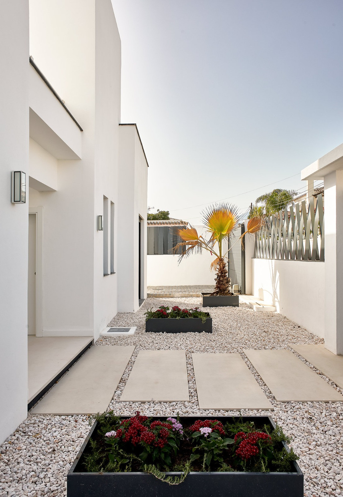 Exterior side path with white gravel, large paving stones, and dark planters with flowers and a small palm.