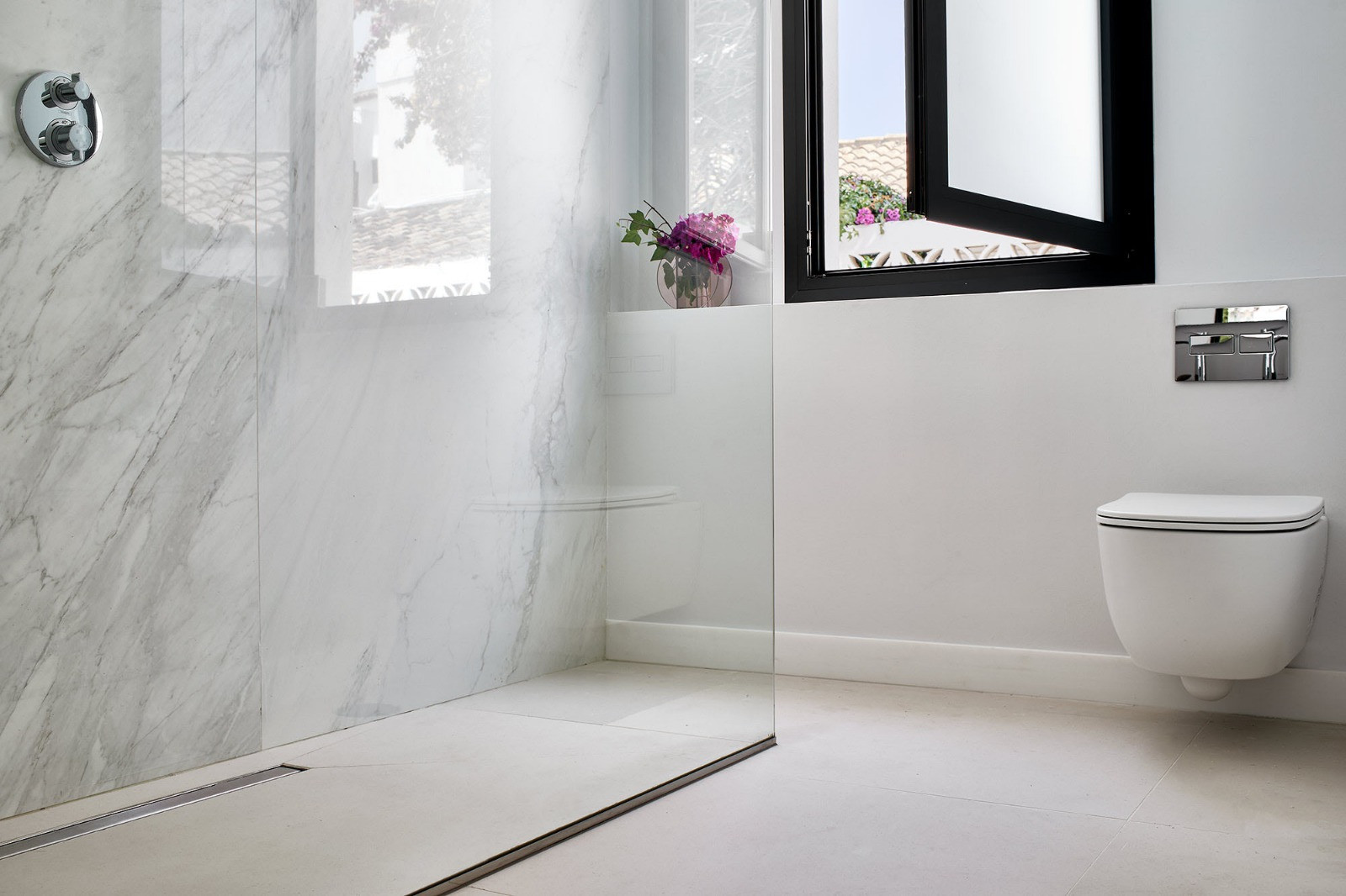 Modern bathroom featuring a wall-mounted toilet, glass shower enclosure, and an open window with white shutters.