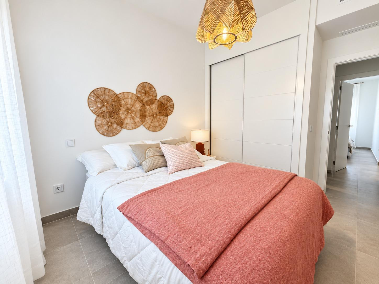Bedroom with a bed featuring a terracotta throw, wicker wall decor, a wicker ceiling lamp, and white sliding wardrobe.