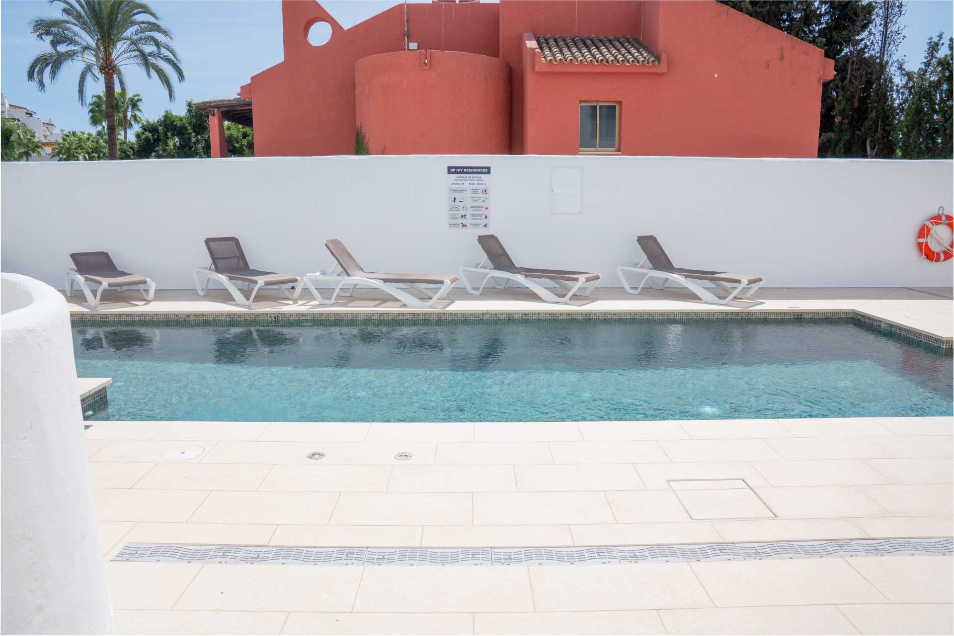 Outdoor swimming pool with five lounge chairs and a white wall, backed by a red building and palm trees.