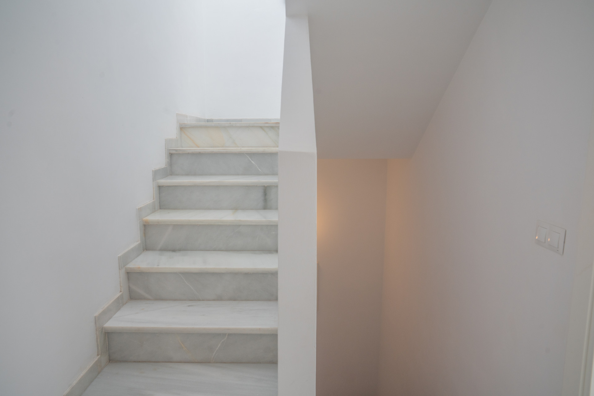 Upward view of a white marble staircase with white walls and a light switch on the right.