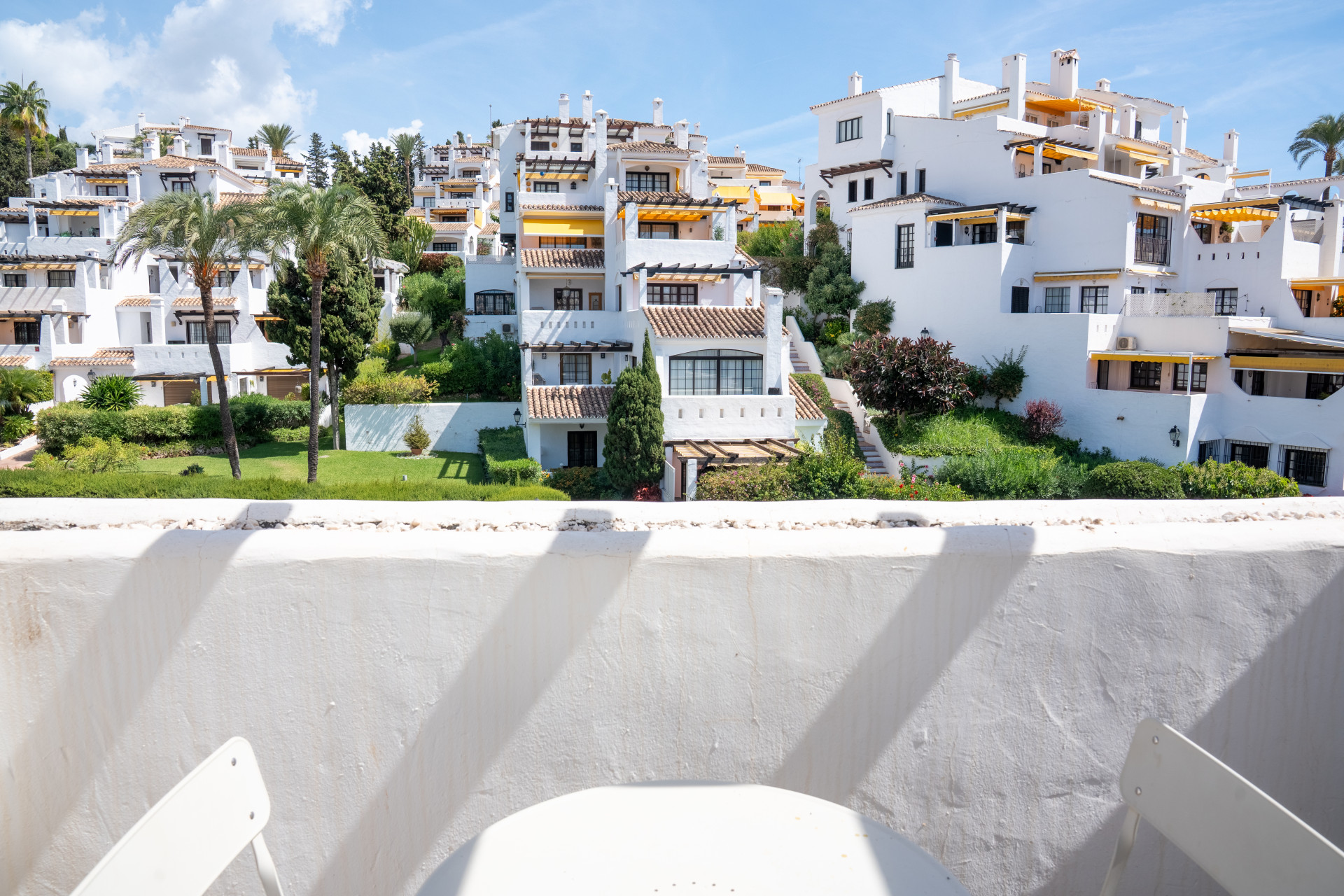 View from a balcony showing white hillside buildings, green landscaping, and palm trees under a blue sky.