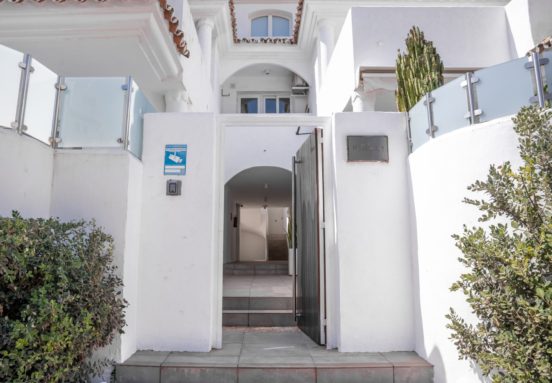 White building exterior with an open door leading to a hallway, flanked by green bushes and glass railings.