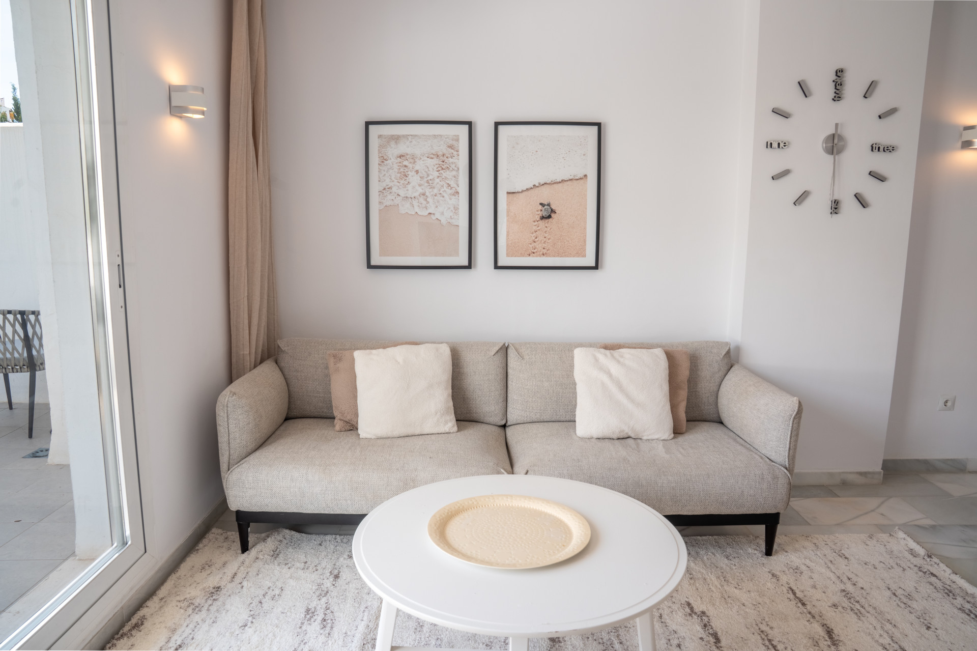 A neutral-toned sofa with pillows and a round coffee table in a living room, with art and a clock on the wall.