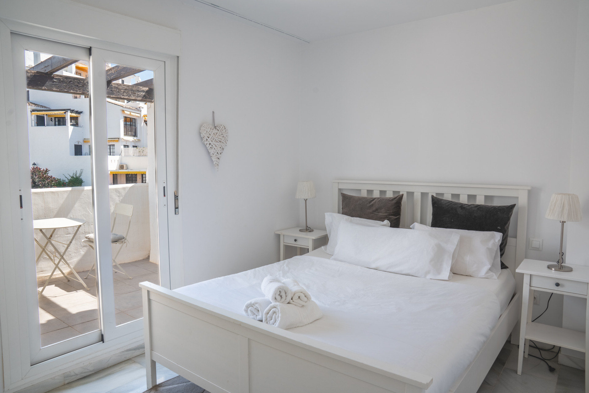 Bedroom with white bed and sliding glass door to a balcony. Two lamps and a heart wall decor visible.