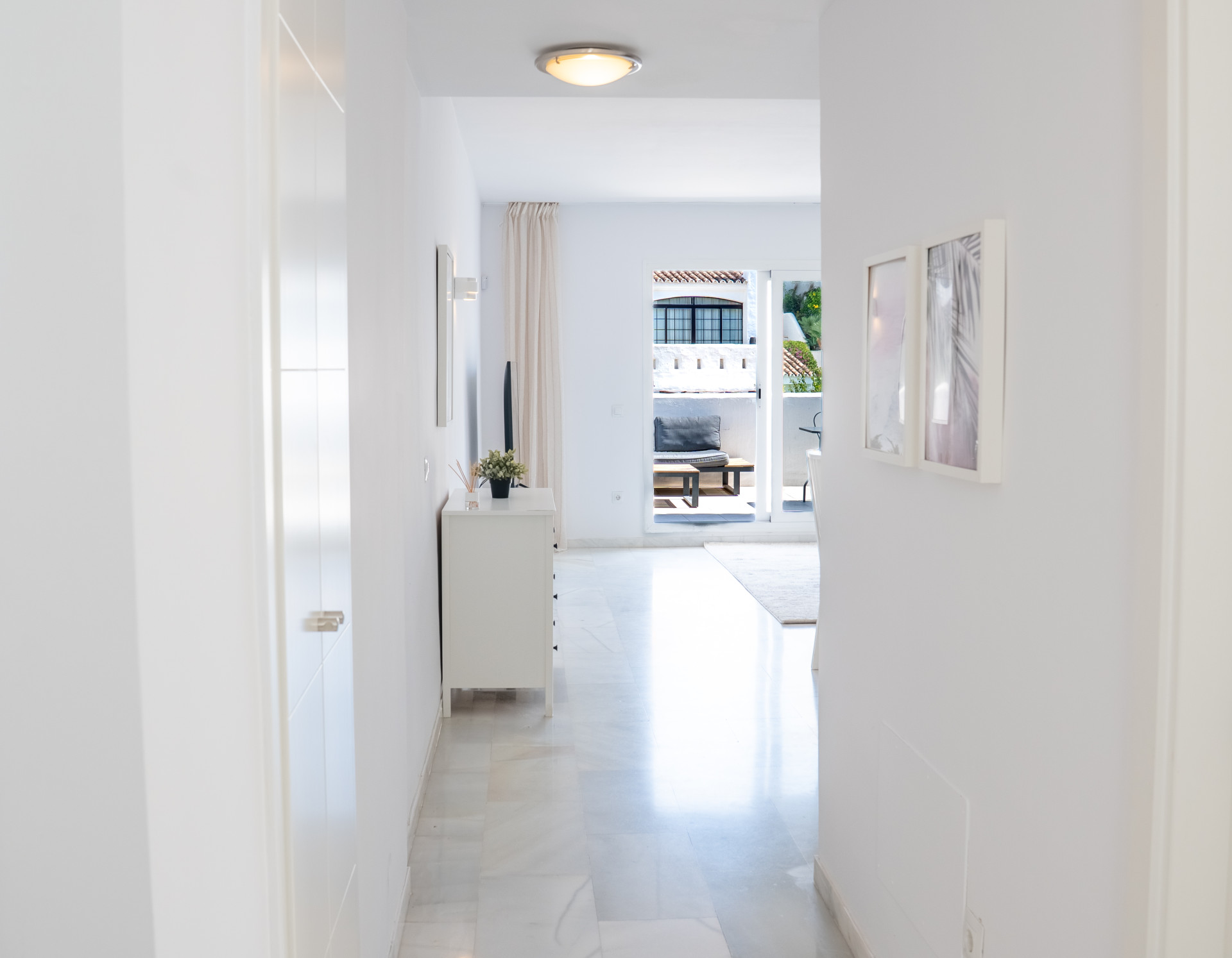 White hallway leading to a bright living area with a glimpse of an outdoor balcony. Marble floor tiles visible.