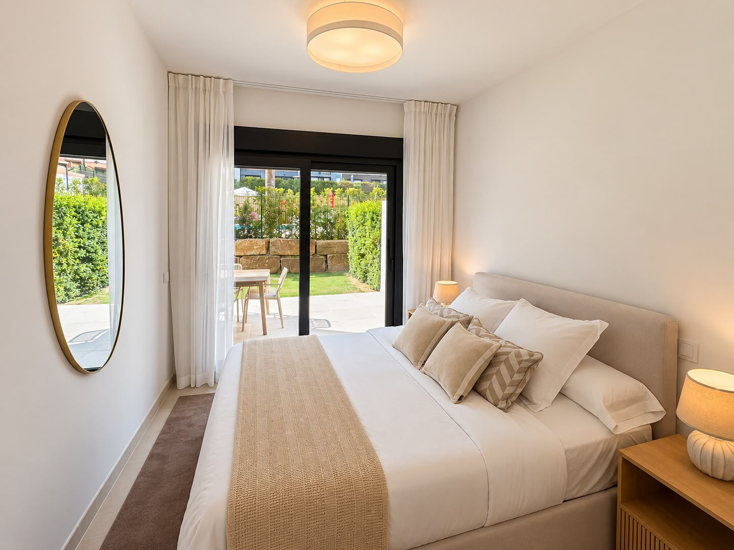 Bedroom with a large bed, two nightstands, a round mirror, and glass doors leading to a patio and garden.