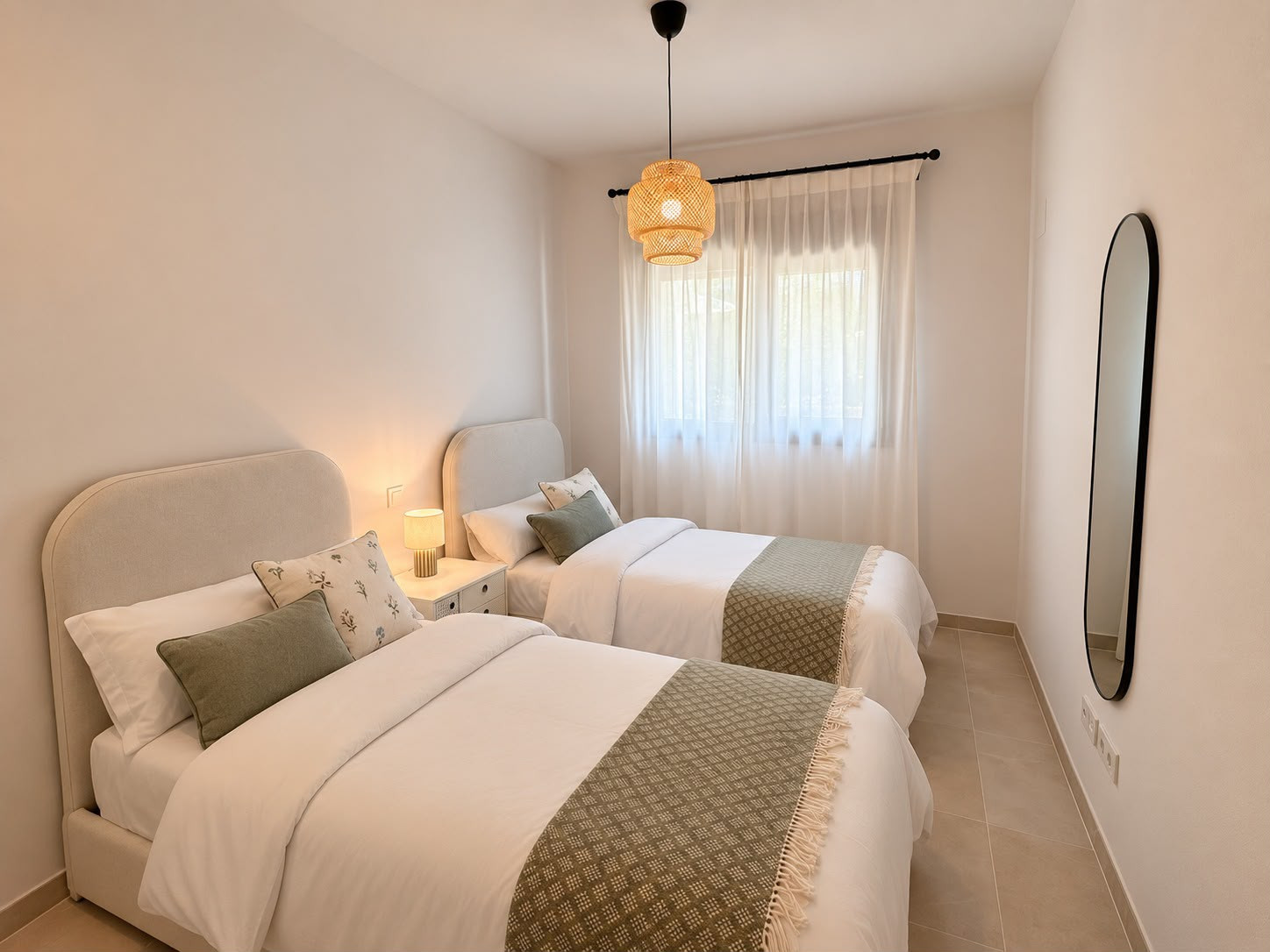 Bedroom with two single beds, central pendant light, window with sheer curtains, and a tall mirror.