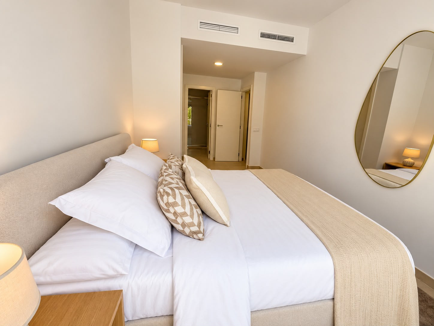 Bedroom with a beige upholstered bed, white linens, two nightstands, and an oval mirror, facing an open doorway.