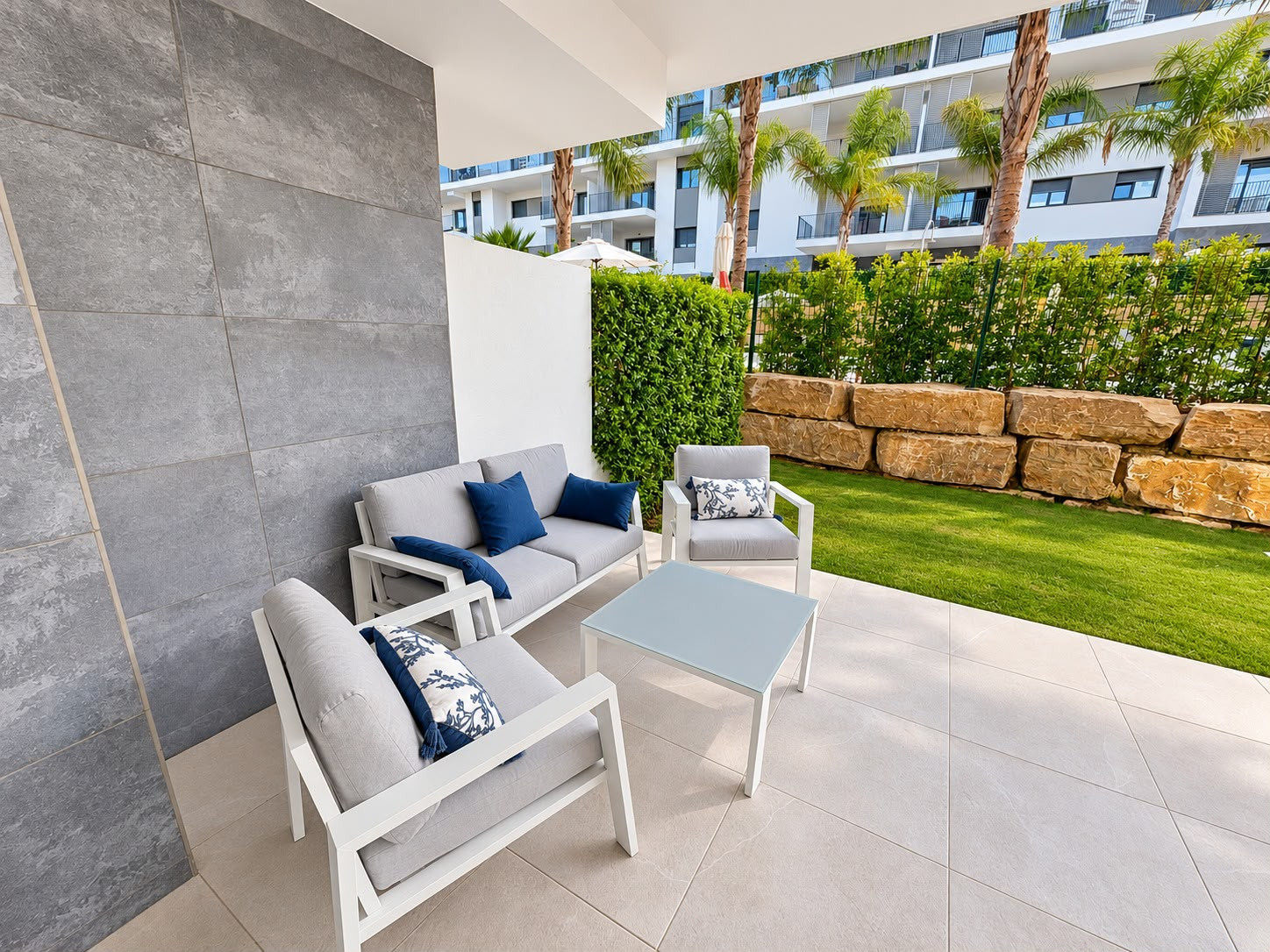 Outdoor patio with light grey seating, blue cushions, a small table, and a manicured lawn.