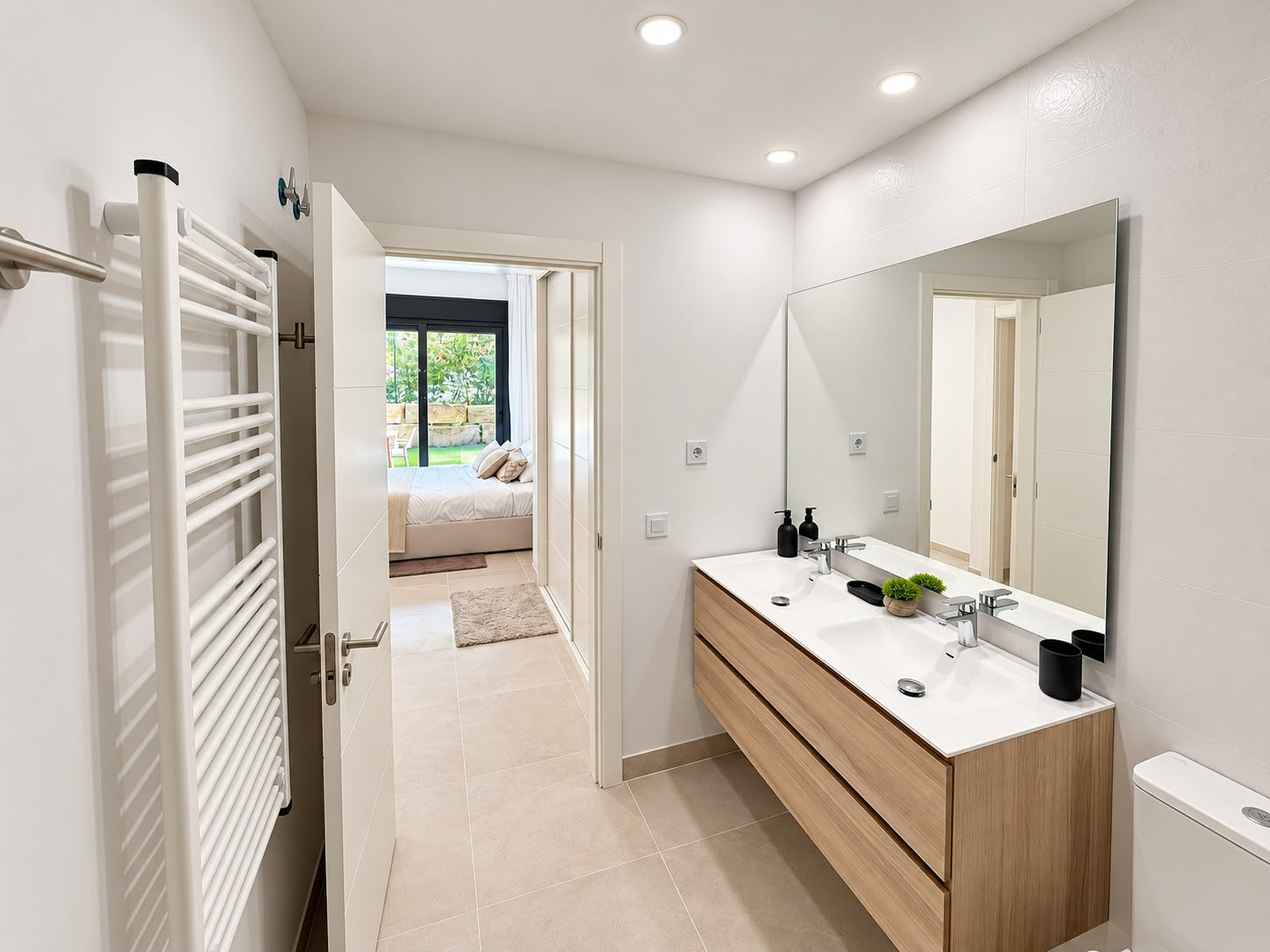 Bathroom with a dual sink vanity, large mirror, heated towel rack, and an open doorway to a bedroom.