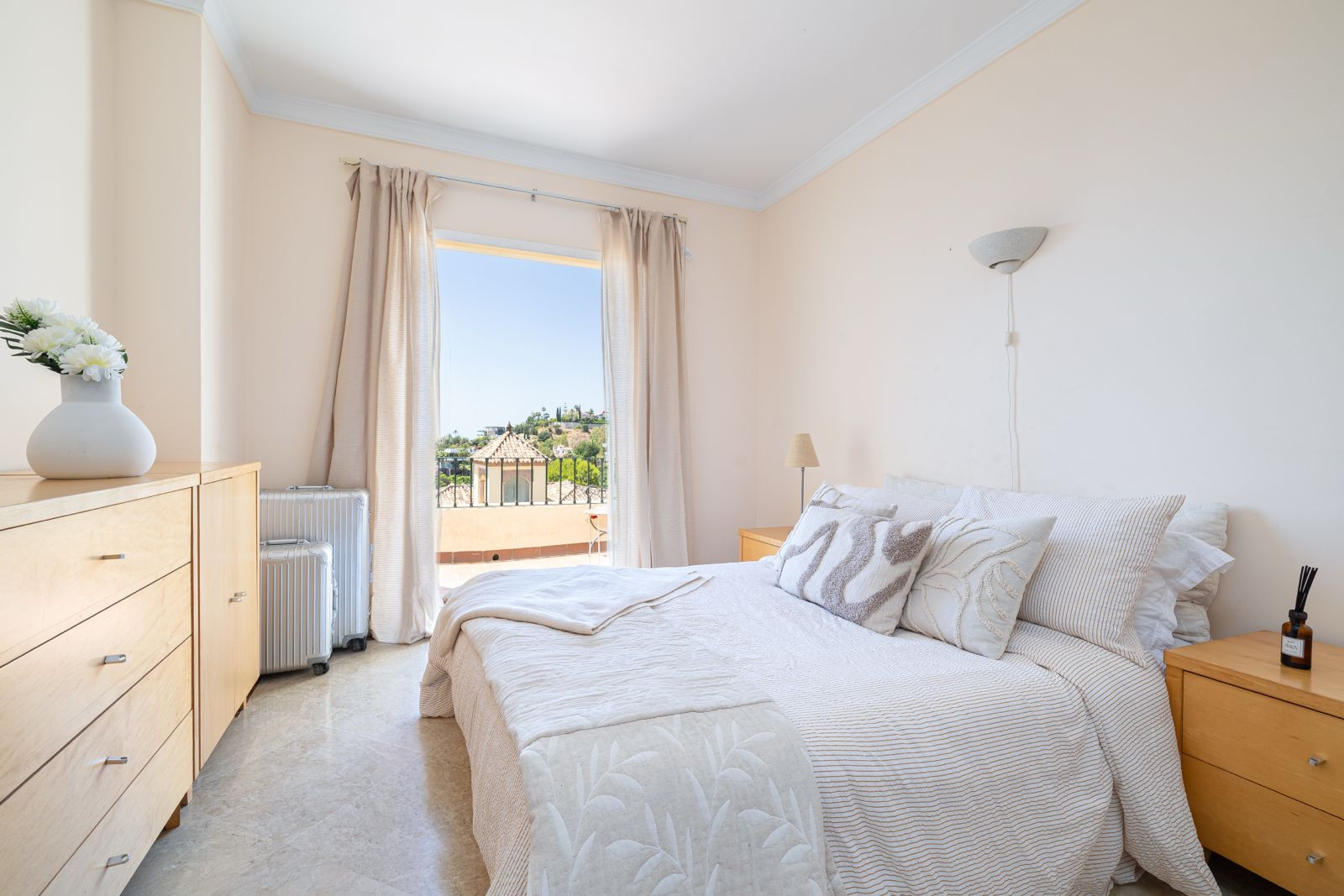 Cozy bedroom with a large window, dresser, and luggage next to the bed gleaming in natural light.