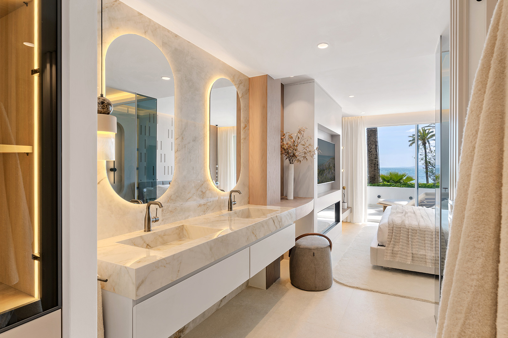 Bathroom with double vanity sinks, illuminated mirrors, and a clear view into the bedroom and balcony.