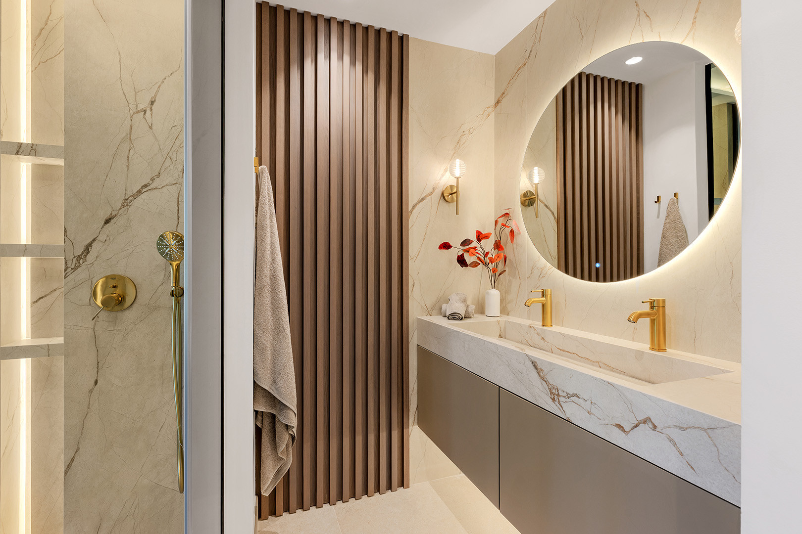 Bathroom with a double vanity, round lighted mirror, gold fixtures, and a shower with marble accents.